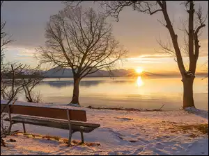Bezlistne drzewa i ławka na śniegu nad jeziorem z widokiem na zachód słońca nad górami